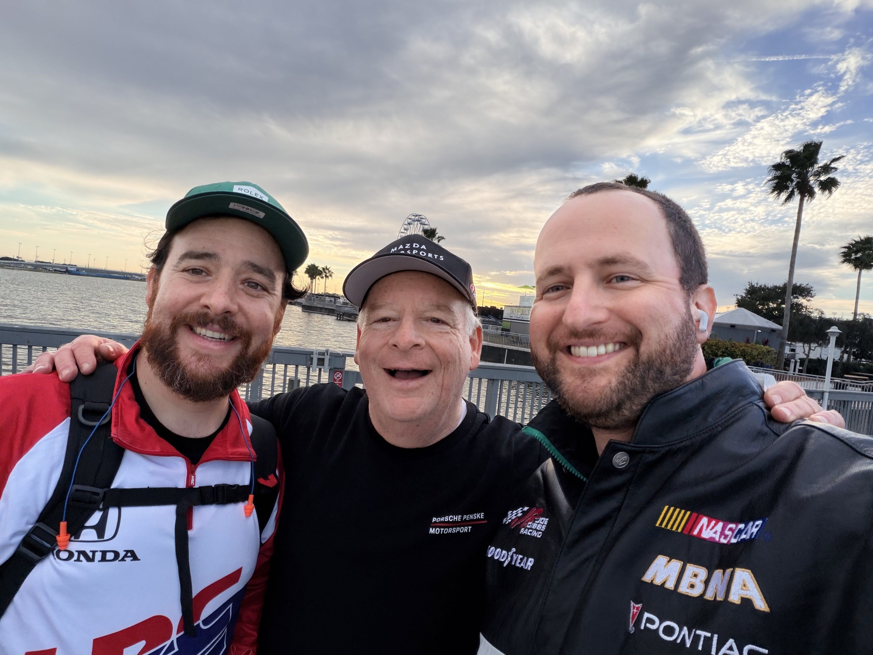 Three men in racing gear smiling for a selfie on the pier over Lake Lloyd at Daytona International Speedway